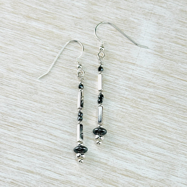 long silver and grey earrings hanging from a silver hook. The pattern, from top to bottom is a small grey round bead, a short shiny silver square rod, 2 small grey round beads, another square rod, a flat round glossy grey bead wider than the others and a round silver bead.