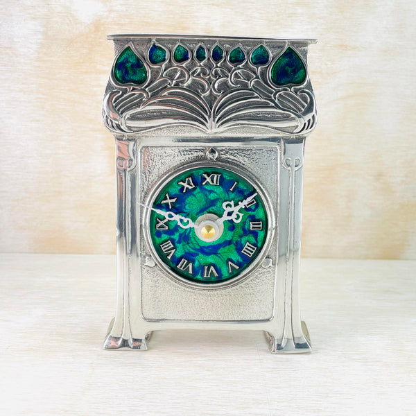 Shiny pewter clock in a rectangular shape. The top third has branch like decorations with eight blue enamel  leaves at the top - different sizes. The clock face is central to the bottom part of the clock, is round with a blue/green face, silver coloured roman numeral numbers and ornate silver coloured hands. A column down both sides join to the slightly curved feet.