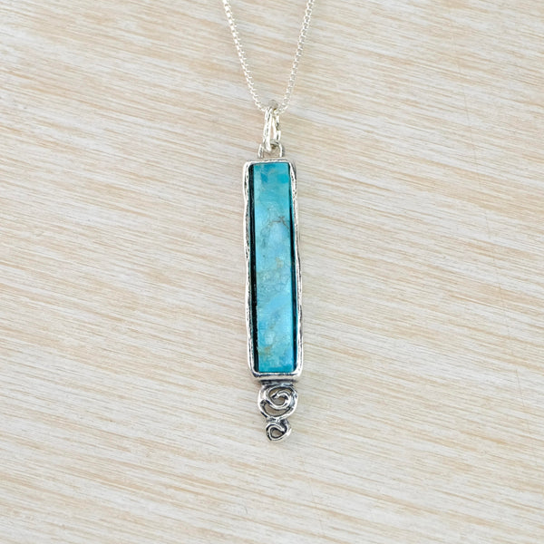 Long slim slender turquoise stone is set within a silver frame with a  hammered silver bale and a small silver swirl at the bottom.