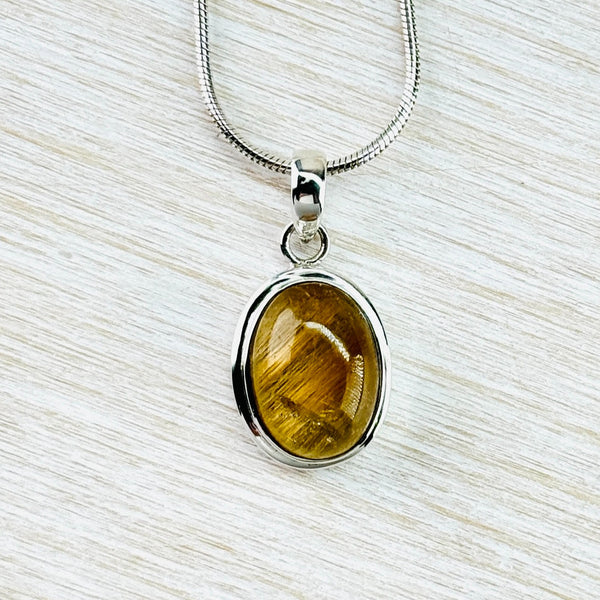 Oval shaped stone simply surrounded in silver. The stone is golden colour with delicate stripes across the stone, like golden strands. It hangs off a silver chain.
