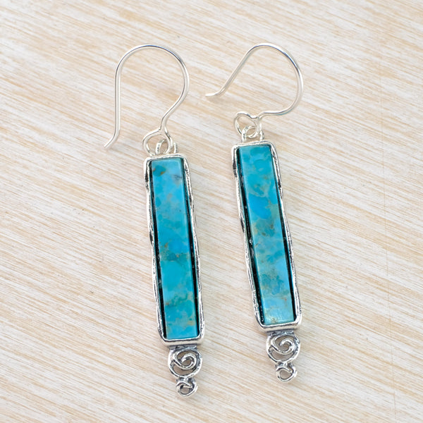 A long rectangular turquoise stone is framed by silver with a decorative scroll at the bottom. Hanging from a silver hook.