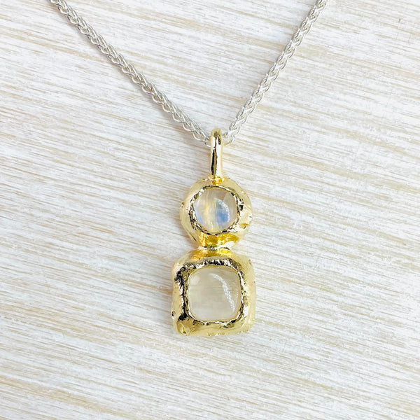 A smaller round moonstone sits above a slightly larger square one. The top stone has more blue reflections. Both are set in textured gold plating in a deep frame. Hanging from a silver twisted chain.