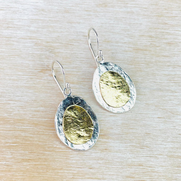 Two slightly irregular shaped ovals. A gold coloured one is laid on top of  slightly larger silver one. Both are textured, to look a bit like crumpled tin foil, and hang from a silver hook.