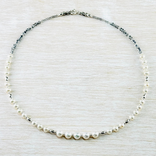 A beaded necklace with beads all the way around. It has a symetrical design. At the front are four larger round  pearl beads , with little silver spacers between them. Then (on each side) a silver bead, three smaller pearl beads, a silver bead, four smaller pearl beads, a silver spacer, two same size pearls, a spacer, four pearl beads, then small grey crystal beads and small cut rainbow moonstone beads. Finished with a round spring catch.