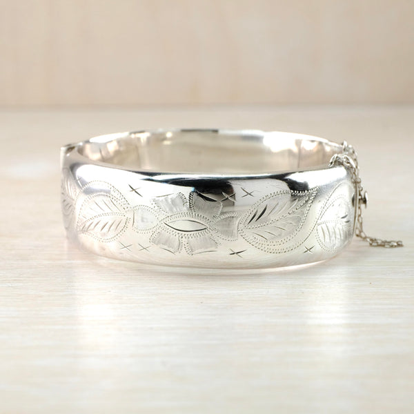 Shiny silver bangle with a safety chain on one side. It has a delicate engraved design of a central flower and leaves with plain high polished silver top and bottom.