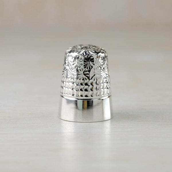 Shiny silver thimble decorated in 3 ways. The top has star shapes, or possibly star shaped flowers, the middle is a repeated waffle like design and the bottom third is plain and high polished.