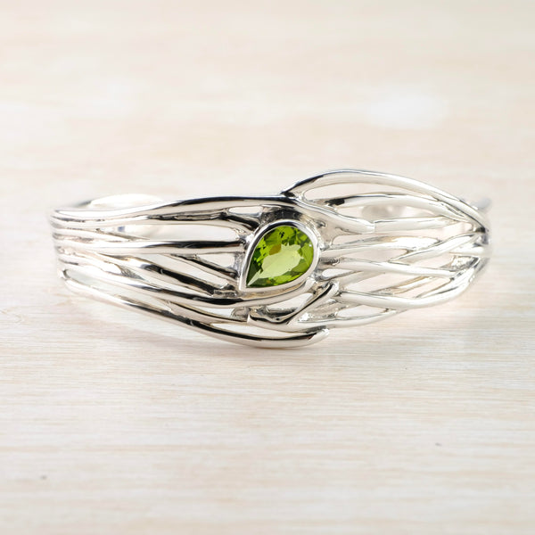 A light green, cut, sparkly tear drop shaped stone is set centrally at the front of the bangle. It points to bottom left side rather than straight down. Eight strands of shiny silver form the bracelet at the front. overlaying each other.