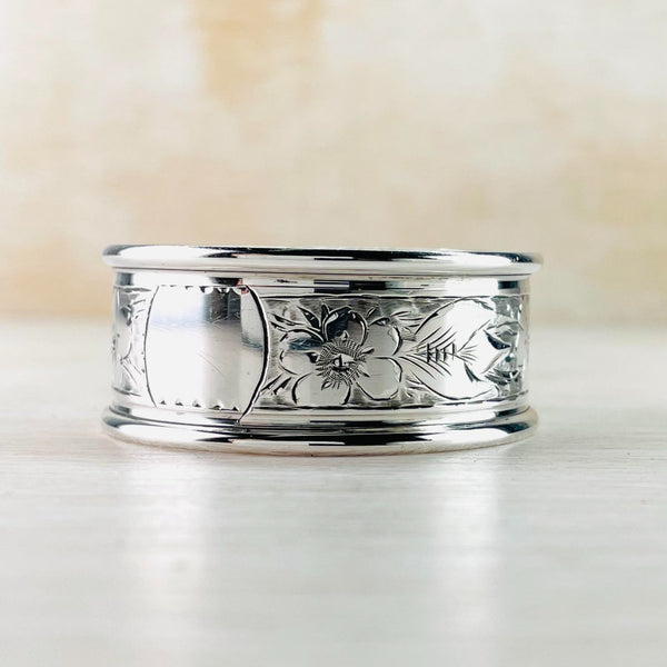 Shiny silver with two plain rims. The inner section is engraved  with 5 petalled flowers, probably forget me nots , and leaves all the way around except for a plain circle for engraving.    