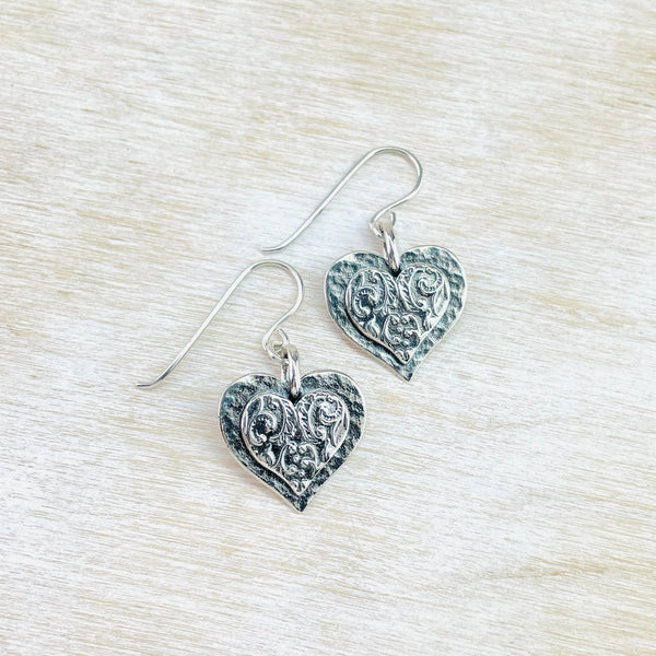 pair of earrings made up of two hearts, one on top of the other. Both hearts are textured silver, the top one, slightly smaller gas scroll decorations in it. Simply hanging from a hook.
