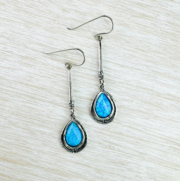 Tear drop shaped vibrant bright blue opal stones are set within a double silver  frame. One frame is polished silver, the second is darker silver with delicate swirl decorations. This is hanging from a slender silver stalk with a double stripe at the bottom. All hanging from a simple silver hook.