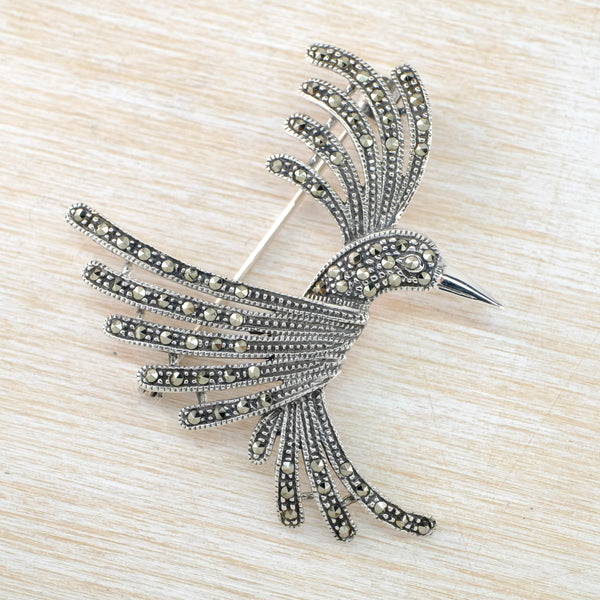 A humming bird in flight viewed from the side. Each wing has five feathers in silver with shiny dark grey little marcasite stones set along them. The tail has similar four feathers. It has a long shiny beak and a pretty little eye.