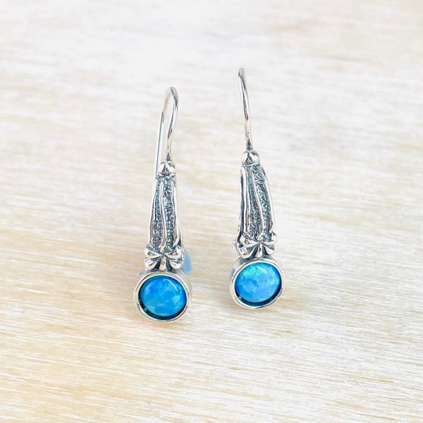 Bright blue round opals are set at the bottom of a long almost triangular piece of silver. The silver has an upside down flower flower on a stalk decoration and is attached to a hook at the narrower end.