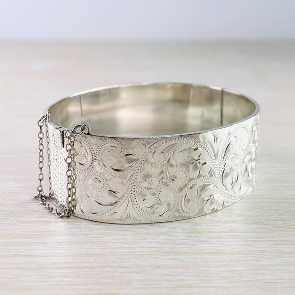 A wide bright silver bangle decorated in a fleur de lys design some decorated by lots of little silver dots. The whole of one side is covered with this swirly decoration, the other is plain.