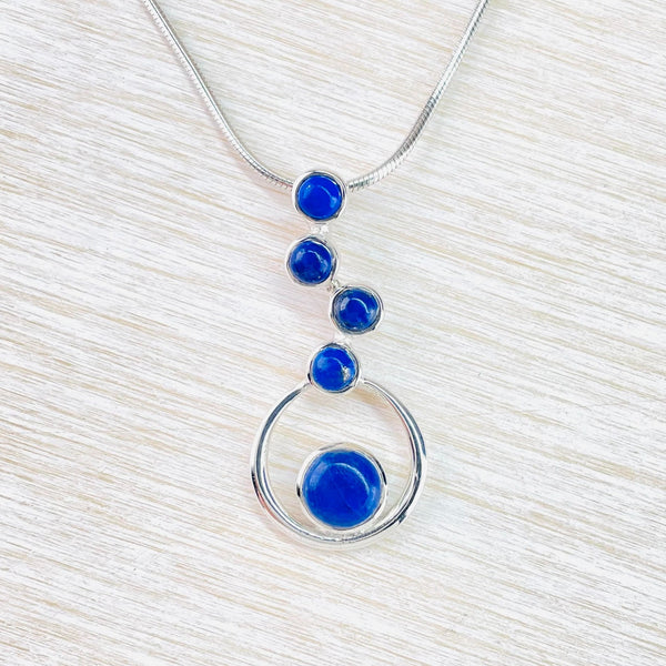 Four circular deep blue lapis stones , set in silver, are in a slightly staggered vertical display. Below these is a silver hoop with a larger round lapis stone, also set in silver, sitting at the bottom but still inside the hoop.