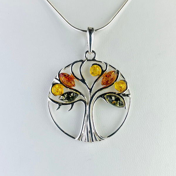 Silver and amber pendant with an overall circle shape. Wiuthin the silver circle sits a tree - textured trunk at the bottom, slender branches reaching the top. There are 7 silver branch with amber leaves nestling amongst them. There are seven leaves in total, some round some diamond shaped, in 3 different shades of amber - green, golden and orange.