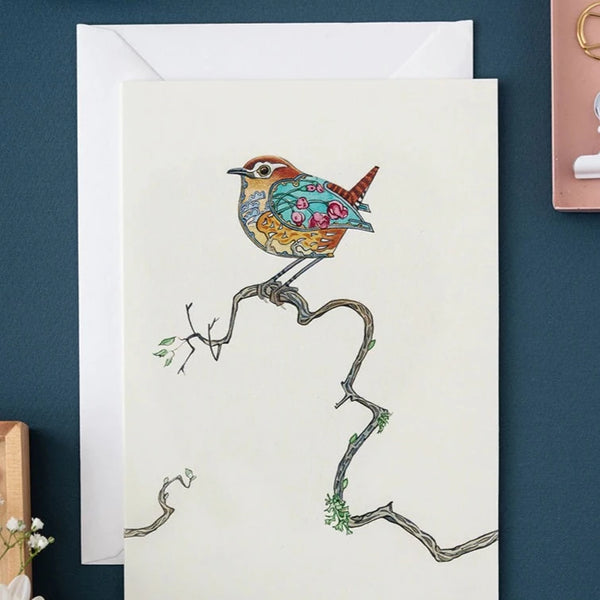 A little wren sits on a wavy branch with with a few leaf buds on it. The wren is decorated  with pink flowers on a blue background on it's wing and tree designs in shades of brown on it's tummy. Bright eyes and a little pointed beak.