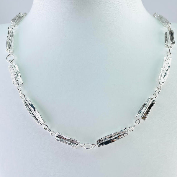 This heavy silver necklace is formed of 15 links. Each link is polished textured silver in a rectangular shape. The texture looks a little like the bark of a tree. 7 of the links are slightly longer than the other eight and they are arranged alternately, linked with plain silver jump rings.