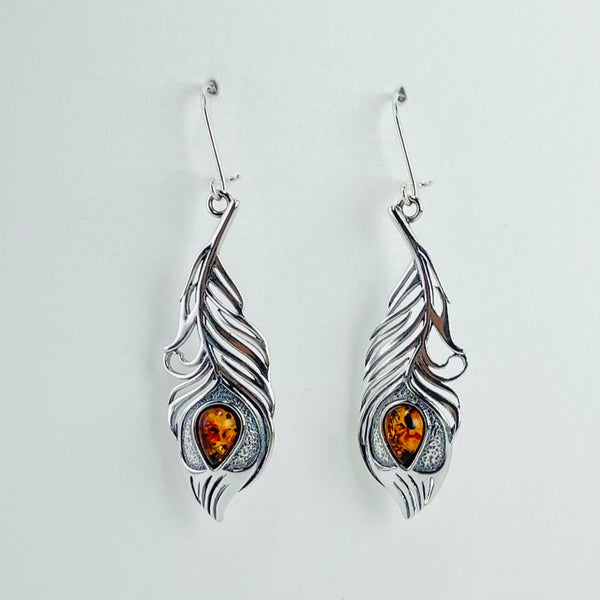 Earrings in a peacock feather design. They are a mirror image of each other. The majority of the feather is in silver with some open parts. The shape is exactly like a peacock feather with the eye being represented by a tear drop piece of golden coloured amber. Hanging from a silver hook.