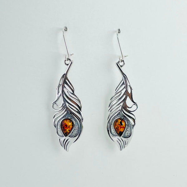 Earrings in a peacock feather design. They are a mirror image of each other. The majority of the feather is in silver with some open parts. The shape is exactly like a peacock feather with the eye being represented by a tear drop piece of golden coloured amber. Hanging from a silver hook.