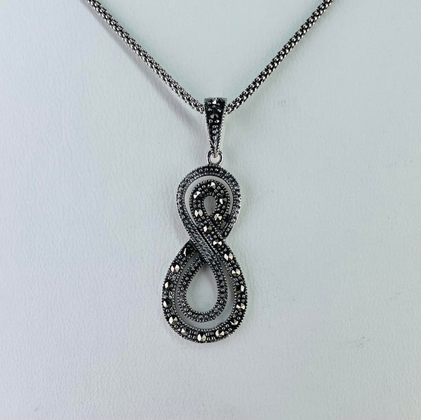 The pendant is in the shape of an eight, with the top half being a little smaller than the bottom half. the shape is formed of two strands, a narrower inside at the bottom, outside at the top as they twist in the centre. The narrower strand is set with smaller marcasite stones. it hangs on a bail set with marcasite and comes on an oxidized chain.