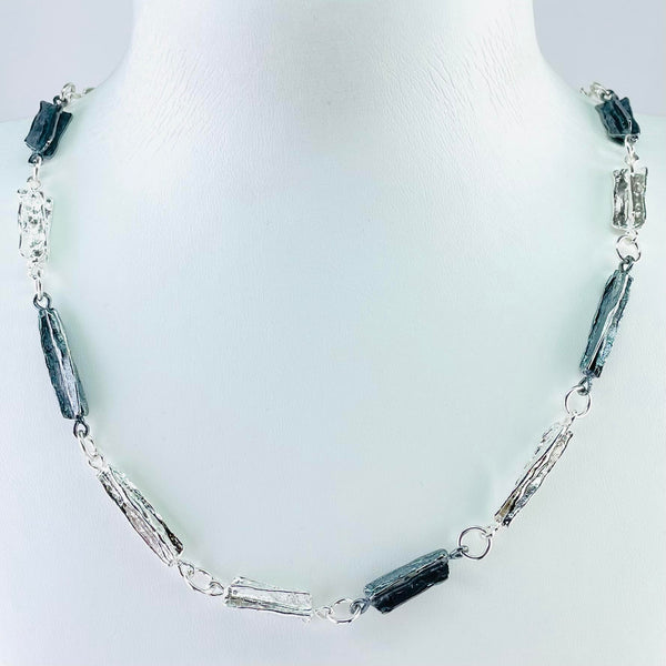 This heavy silver necklace is formed of 15 links. Each link is  textured silver in a rectangular shape, some are bright silver, some oxidized. The texture looks a little like the bark of a tree. 7 of the links are slightly longer than the other eight and they are arranged alternately, linked with plain silver jump rings. The oxidized links are randomly placed along the necklace.