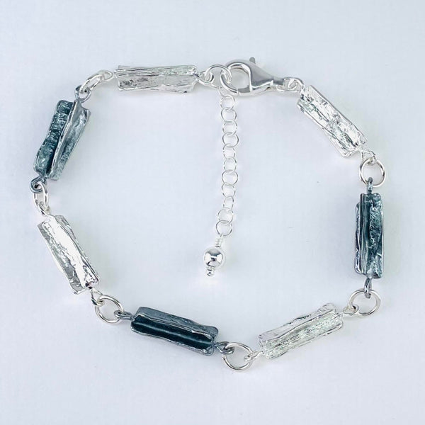 A bracelet made up of seven rectangular links  . They are all textured silver, a texture that looks a little like the bark of the tree. Four of the links are high polished, three are oxidized, so look dark grey. They are linked by round jump rings and finished with a heavy lobster catch. A plain silver chain hangs at the end of the bracelet with a little silver bead at the end ( so it can be worn a little longer)