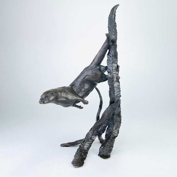 A sculpture of an otter swimming through water reeds. The otter is angled downwards, front feet tucked in towards the body, back feet stretched behind by the side of a tall reed. The tail is at a steep angle upwards, head streamlined with eyes and nose visible. There is one tall reed  pointing upwards and smaller ones at the base.