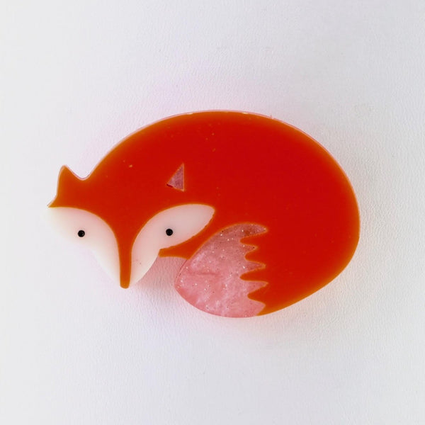A simple fox shape, head at one end, curved body shape and a tail almost tucked in under his chin. It almost forms a circle. Fox orange in colour, the face has white cheeks, little black dot eyes and pointed ears. There is white shading at the end of his tail.                                                            