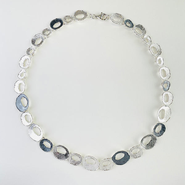 A necklace formed of off circle linkes with an off circle cut out.All are textured, some are dark oxidizes silver, some brighter.