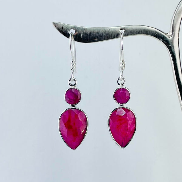 Double drop ruby quartz earrings. The stones are a deep pinkish red and have been faceted. The top stones are smaller than the bottom ones and are circular, simply set in silver. The lower stones are pear shape, wider than the top and longer, also set in silver. The stones hang from the ear wire, connected by a silver ring.