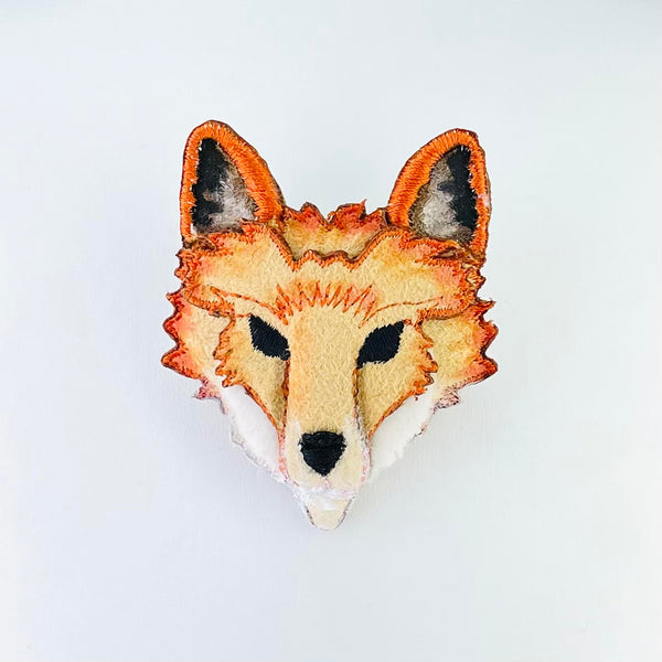 A foxes head with perked up ears - orange edged with black and grey creating the inside of the ears. Black eyes and a black nose set onto a pale orange face, with darker orange shading and white cheeks.
