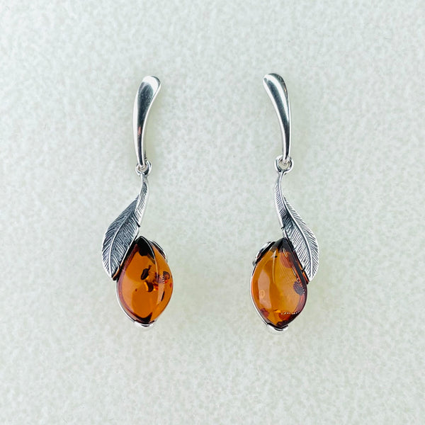Each earring is a curved silver leaf shape, with oxidized silver creating the vein design on the leaf. On the inside of each leaf , from about half way down, a golden coloured piece of amber, with a slightly wavy shape, is attached to run parallel to the leaf and then hang lower. The amber has quite clear, with just a few inclusions reflecting the light. Hanging from a silver stalk with a post and butterfly fitting behind.