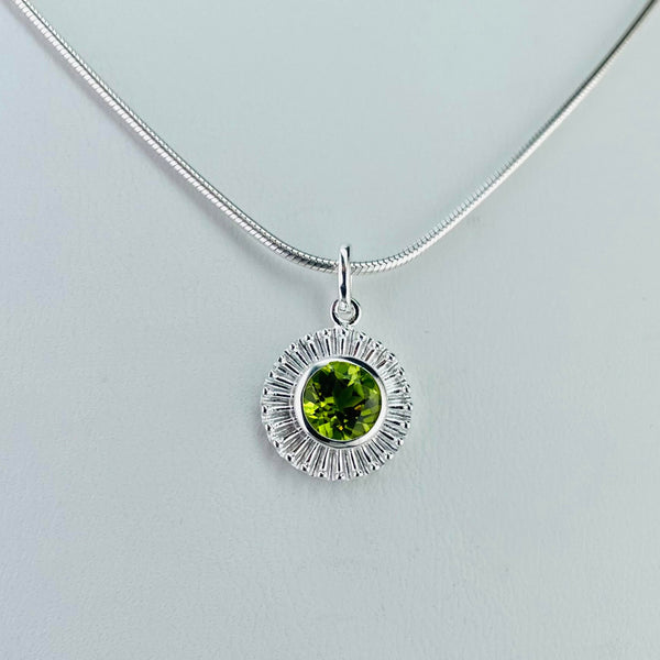 A round bright lime green faceted peridot stone is set within a silver surround formed of lots of closely spaced 'rays' forming a solid surround. There is a plain silver circle between the stone and the rays. The pendant has a simple silver bail and a shiny silver snake chain.