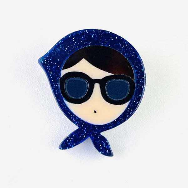 A cream female face forms the centre of this brooch. She has dark hair showing as a fringe with a side parting, is wearing large sunglasses and just has a dot as a mouth. She has a dark blue glittery scarf tied around her head knotted under her chin, a little like the Queen would wear.        