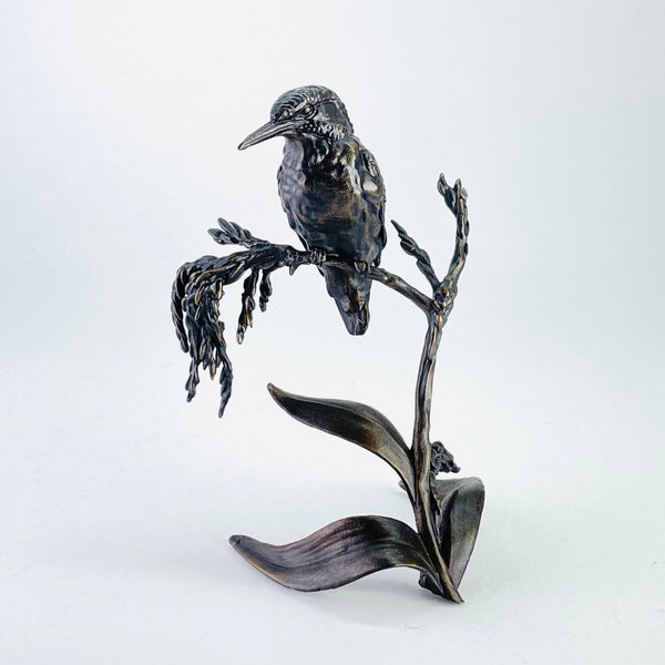 All bronze coloured. A kingfisher is sitting on a water plant. Three leaves are at the base, curling down slightly. Then there is a stalk which forks at the top. One fork has a grassy head with the kingfisher sitting half way along, with its long beak turned to the left and beady eyes.