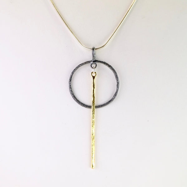 Simply formed of an oxidized circle with a polished gold plated slim stick hanging from the central top of the circle down to about twice its length.
