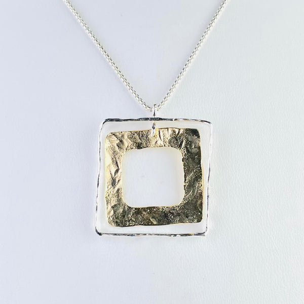                                                         A square pendant on a silver chain. The outer piece of the pendant is a square silver frame, hanging within this so it moves seperately, is a gold square thicker than the silver one, with a textured finish.