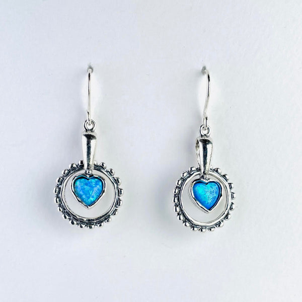 Drop earrings with a patterned silver circle  below a plain silver stalks. Hanging from the stalk within the circle is a bright blue, heart shaped opal surrounded in silver.  