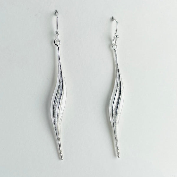 Slim long silver earrings in a delicate wavy design. Each earring has a central stripe which is a little darker. They are narrower at the top and bottom and are attached to a silver hook.