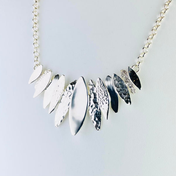 Silver necklace with a central display at the front which is formed of 12 different sized leaf shapes with different textures of silver. The longer shape is in the middle, getting smaller as it goes around the neck.