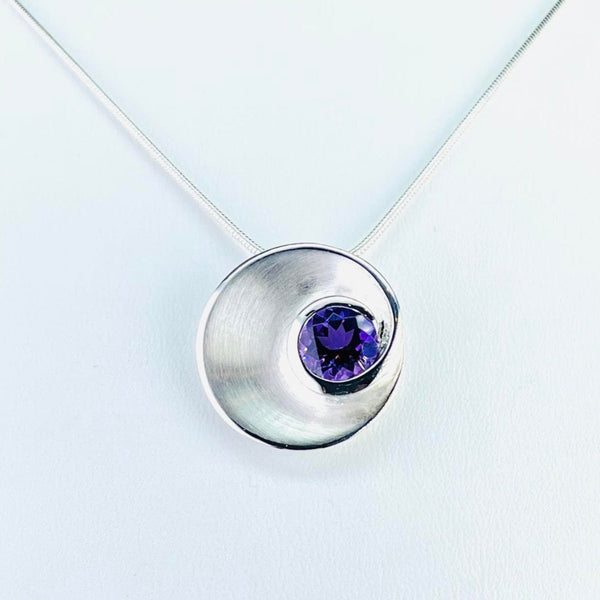 A satin silver circle with high polished edging and a slight spiralling. Set to the right hand side is a dark faceted amethyst within the spiral shape. 