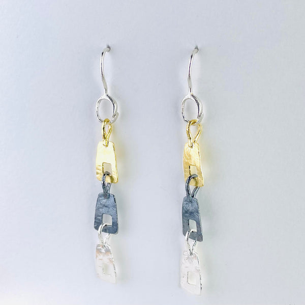 earrings  formed of three  rectangular shapes hanging one below the other. Each has a square cut out. One is gold coloured, one dark silver and one bright silver. All hanging off a hook.