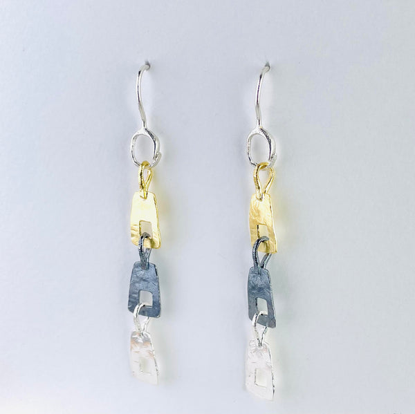 earrings  formed of three  rectangular shapes hanging one below the other. Each has a square cut out. One is gold coloured, one dark silver and one bright silver. All hanging off a hook.