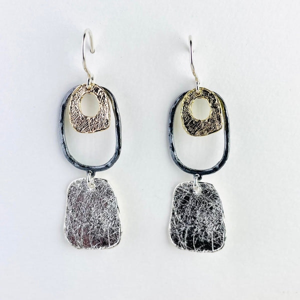 drop earrings in three parts. The top half is a dark silver oblong outline inside which is a smaller gold plated rounded square  with a circular cut out.Below these hangs a solid rounded rectangle.