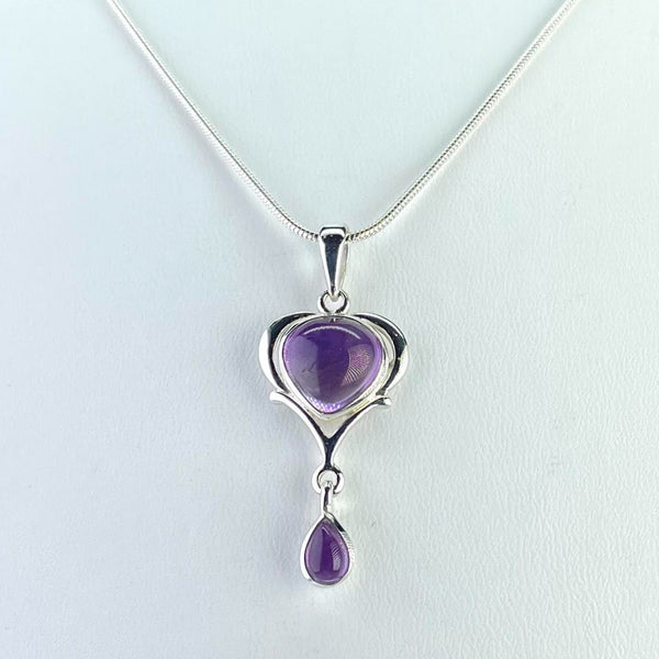 Two amethyst stones hang from one drop. The top one is a soft triangle shape framed in a heart shape silver surround, the bottom one which is much smaller hangs from the top one by a silver loop and is tear drop in shape. Both stones are uncut so have a lovely smooth finish. The wholependant hangs from a polished silver bail. 