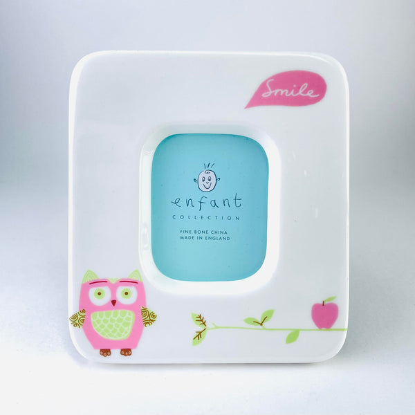  A white china rectangular photo frame - with softened corners. The opening is central, also with softened corners. At the bottom left hand side is a cute little pink owl with large pale green eyes, ears and tummy. To the right is a pink apple on a green twig. On the top right hand corner is a pink speech bubble with the word smile written in white.