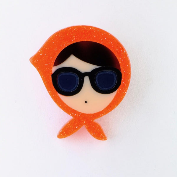 A cream female face forms the basis of this brooch. She has dark hair showing as a fringe with a side parting, is wearing large sunglasses and just has a dot as a mouth. She has an orange glittery scarf tied around her head knotted under her chin, a little like the Queen would wear.