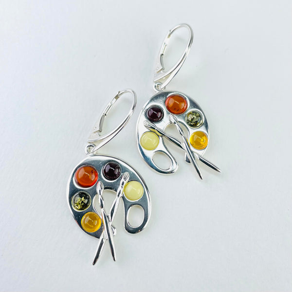 The earrings are in the shape of an artists palette - a broad polished silver horseshoe shape with a hole at one end {where the thumb would go if it was being held.) There are 5 round pieces of amber , that look like the blobs of paint. A golden colour, mossy green,  deep orange, a darker green and cloudy honey colour, Two fine silver paintbrushes are laid over the palettes. They are connected to a lockable silver hook.