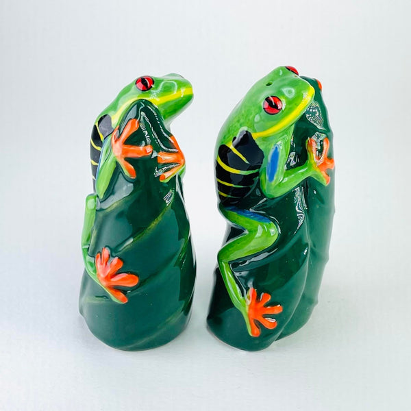 Two bright and cheerful frogs, both clutching on vertically to a dark green pointed leaf. The frogs are holding on tight with splayed bright orange feet. five toes on the back feet, four on the front. The frogs are bright green with a yellow smile, orange and black eyes and black and yellow striped markings on each side.