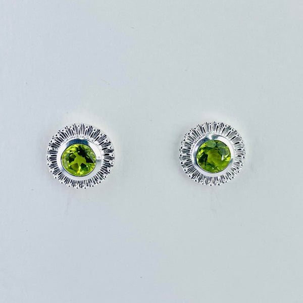 A round bright lime green faceted peridot stone is set within a silver surround formed of lots of closely spaced 'rays' forming a solid surround. There is a plain silver circle between the stone and the rays.